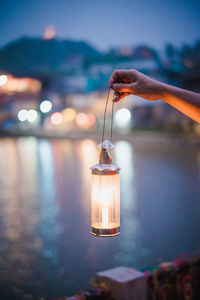 Cropped hand holding illuminated lantern against lake at dusk