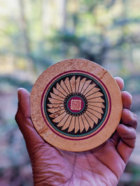 Cropped hand holding patterned wooden circle against trees