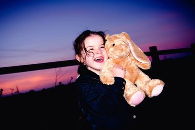 Happy girl with stuffed toy standing on field against sky at dusk