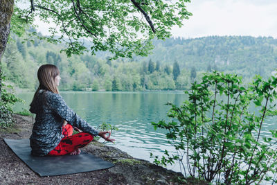 Side view of young woman meditating while sitting by lake against tree in park