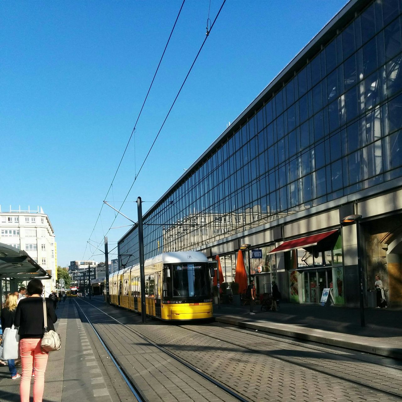 transportation, building exterior, architecture, built structure, railroad track, city, clear sky, public transportation, rail transportation, mode of transport, power line, men, city life, street, railroad station platform, railroad station, walking, the way forward