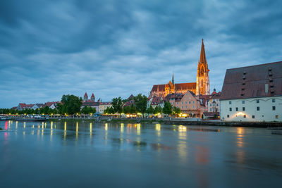 Illuminated buildings by river against cloudy sky