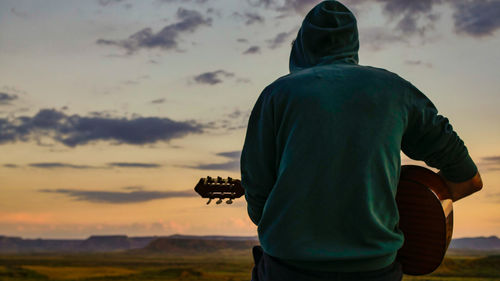 Rear view of man playing guitar against sky during sunset
