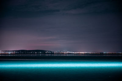Illuminated city by sea against sky at night