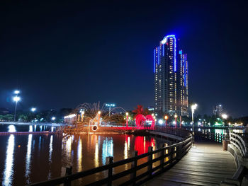 Illuminated modern buildings by river against sky at night