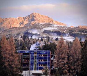 In the foreground between the trees a building behind snow cannons and mountains