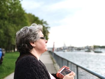 Side view of mature woman holding mobile phone by river in city