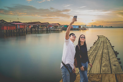 Man taking selfie with woman while standing on pier against sky during sunset