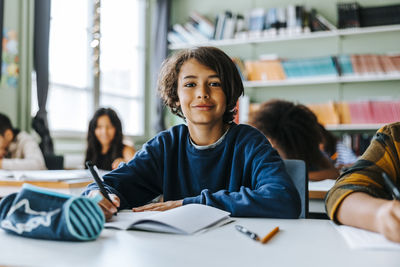 Portrait of smiling boy holding pen while sitting in classroom at elementary school