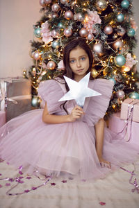 Girl child in a pink dress sits at the christmas tree and holds a star in her hands near her face