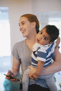 Smiling mother holding mobile phone while carrying son at home