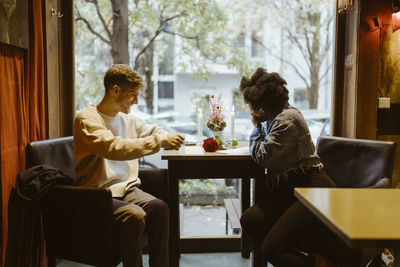 Smiling boyfriend surprising girlfriend with rose while sitting at table in restaurant