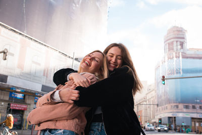 Low angle of smiling female best friends cuddling on street in madrid and enjoying weekend together