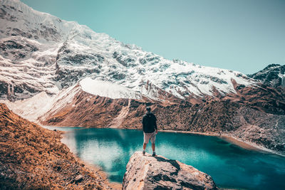 Rear view of man on cliff against lake amidst mountains during winter