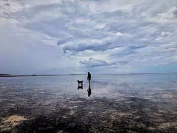 Woman by dog standing at beach against cloudy sky