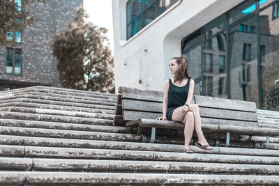 Full length of young woman looking away while sitting on bench in city