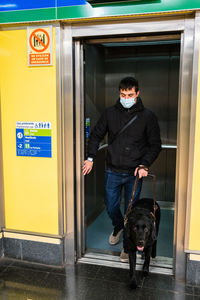 Full body of male with blindness in medical mask calling elevator standing with guide dog