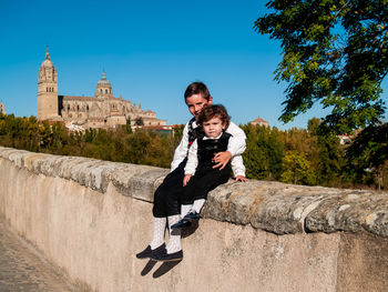 Portrait cute siblings sitting on wall against historic building