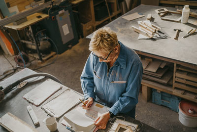 High angle view of senior craftswoman writing on document at workshop