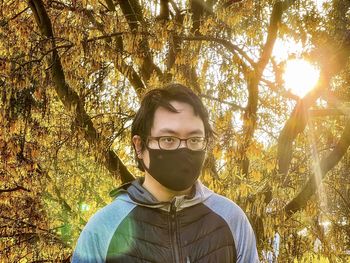 Young asian man in eyeglasses and face mask standing against flowering laburnum tree at sunset.