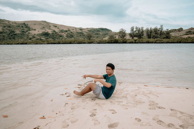 Portrait of young man sitting on shore at beach against cloudy sky