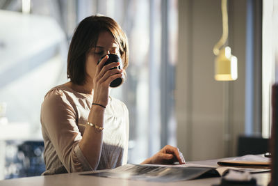 Businesswoman drinking coffee while reading document at desk in office