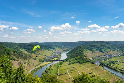 Man flying a green paraglider over beautiful wineries in germany, visible river and forest.