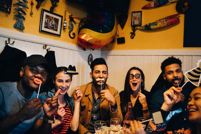 Portrait of cheerful young friends holding props while sitting at restaurant during dinner party