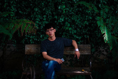 Portrait of young man sitting on bench against plant