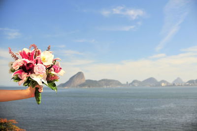 Cropped image of hand holding bouquet against sea at beach