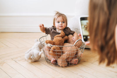 Cute baby girl sitting in basket with soft toys and young mother taking photo on mobile phone 