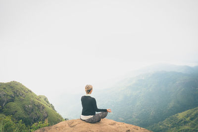 Rear view of young woman sitting on rock against clear sky