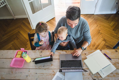 High angle view of man using laptop while sitting with cute son by daughter looking at digital tablet