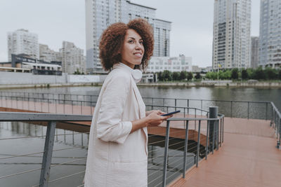 Thoughtful young businesswoman with smart phone standing by railing