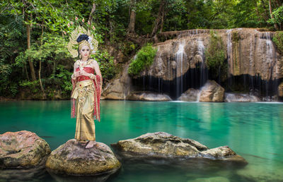Portrait of beautiful young woman wearing crown and traditional clothing while standing on rock against lake at forest