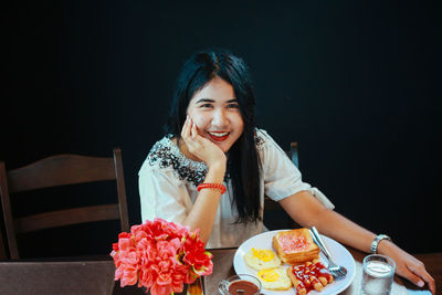 Portrait of smiling woman with breakfast on table sitting against black background