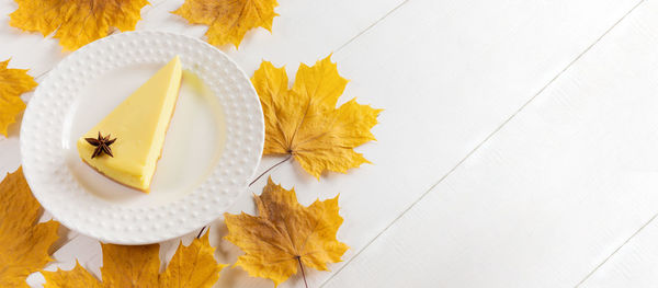 Fall banner with pumpkin pie piece on white plate and yellow maple leaves on white wooden table.