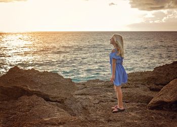 Woman standing on rock at beach against sky during sunset