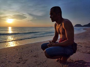 Man sitting on shore at beach against sky during sunset