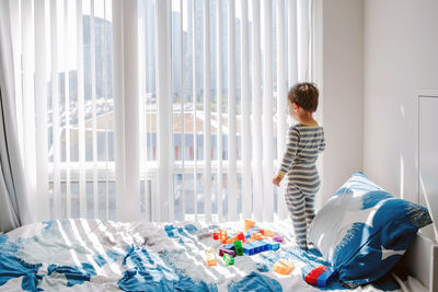 Cute little boy toddler standing on bed in room and looking into window waiting expecting someone.