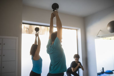 Side view of fit man and woman doing exercises with heavy kettlebells in raised hands during workout in fitness center