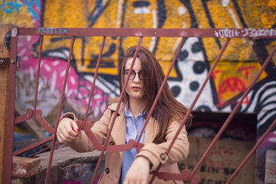 Portrait of young woman standing behind metal gate against graffiti wall