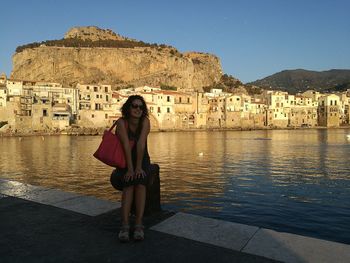 Full length of young woman sitting on bollard at pier by river against buildings