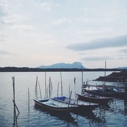 Boats moored by wooden posts in lake against cloudy sky