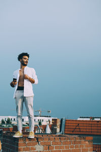 Full length of thoughtful man with smart phone standing on terrace against clear sky