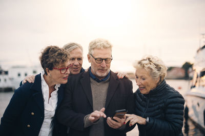 Senior male and female friends using smart phone while standing at harbor