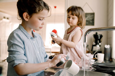 Girl washing mug while sister playing with cleaning brush at kitchen counter