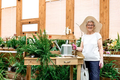 Friendly senior lady with watering can smiling and looking at camera while standing near table in lumber hothouse