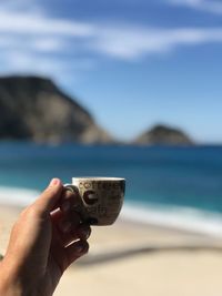 Cropped hand of person holding coffee cup against sea and sky