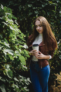 Beautiful young woman holding coffee cup while standing amidst plants